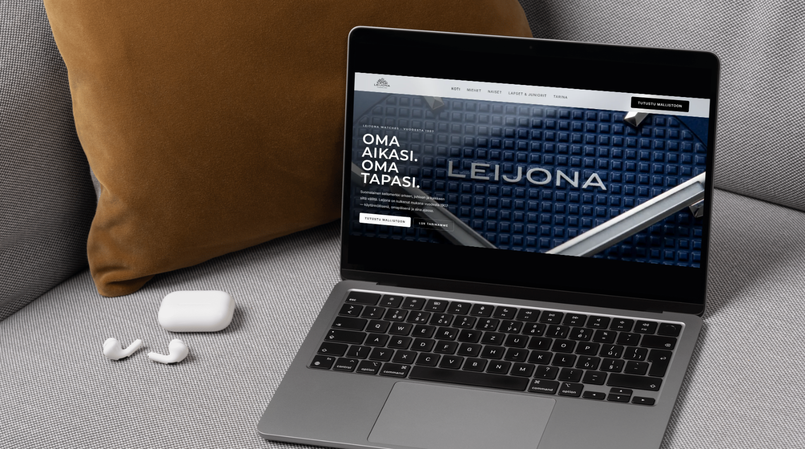 Leijona watch website displayed on a laptop on a grey sofa beside a rust-coloured cushion and white earbuds