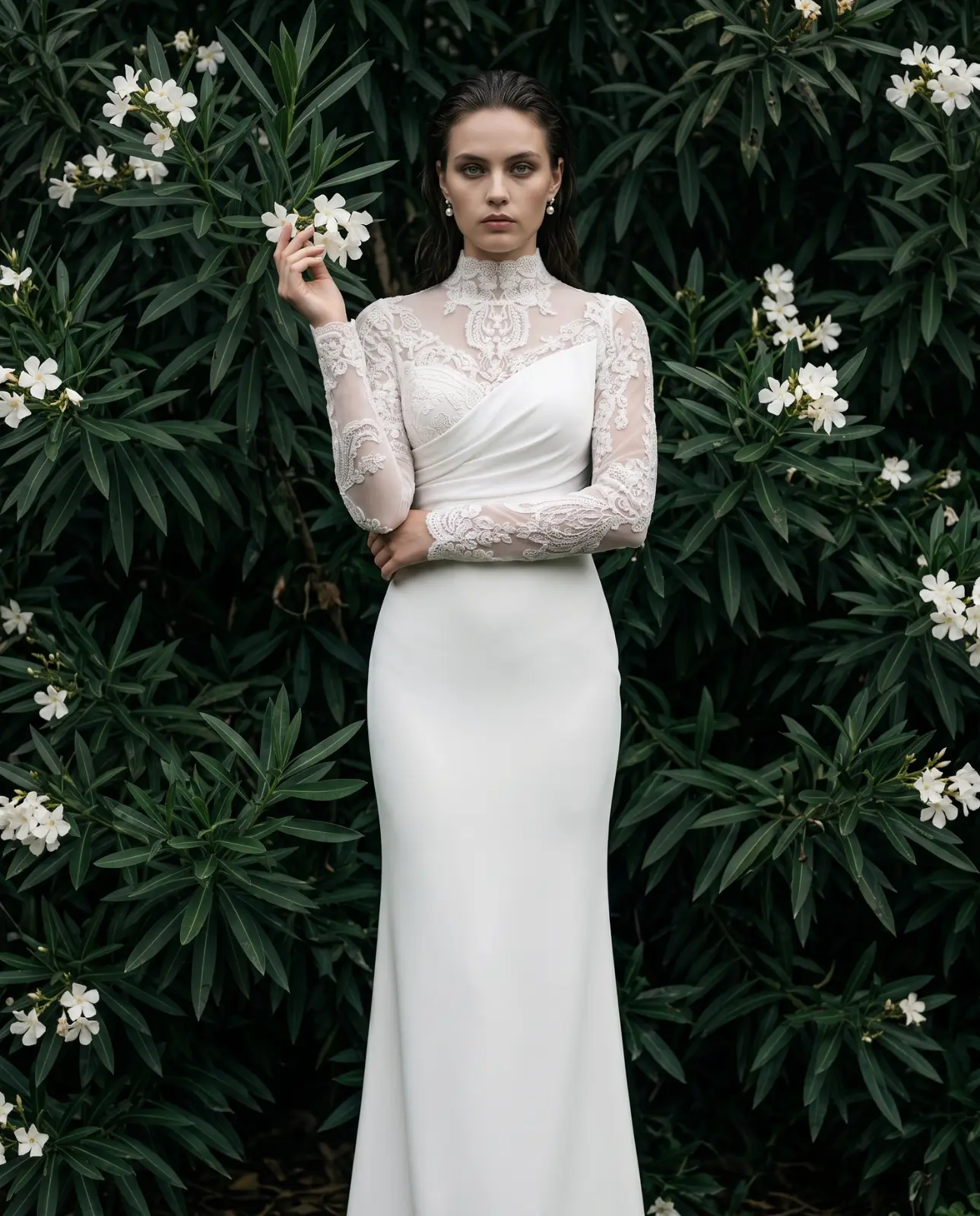 Bride in a lace and satin gown standing before dark flowering foliage